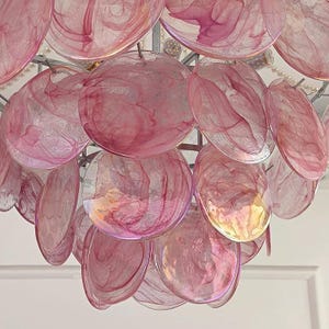 May include: A close-up of a decorative chandelier featuring numerous pink, translucent, shell-shaped elements. The elements are arranged in layers and suspended from a silver-colored frame. The shells have a marbled appearance with varying shades of pink.