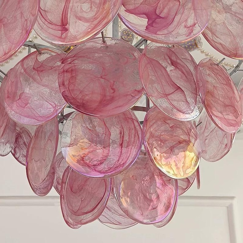 May include: A close-up of a decorative chandelier featuring numerous pink, translucent, shell-shaped elements. The elements are arranged in layers and suspended from a silver-colored frame. The shells have a marbled appearance with varying shades of pink.