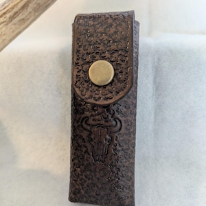 May include: A brown leather case with a textured surface and a brass button closure. The case features an embossed bull skull design. The case is rectangular and appears to be designed to hold a small item.