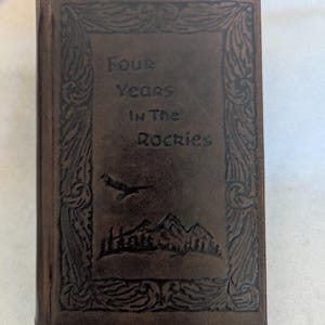May include: A brown leather-bound book titled "Four Years In The Rockies." The cover features an embossed design with an eagle, mountains, and decorative borders. The book appears to be a journal or a memoir.