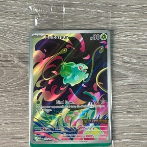 Könnte beinhalten: Pokémon-Sammelkarte mit Bulbasaur-Illustration in Grün und Blau. Der Hintergrund ist holografisch. Text enthält "Bind Down" und "MEGA EVOLUTION". Die Karte befindet sich in einer Schutzhülle.