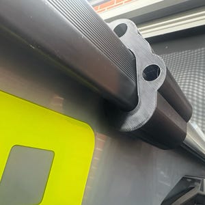Peut inclure: Un support de montage noir imprimé en 3D avec un design profilé à plusieurs trous, fixé sur un rail horizontal noir. Le support présente des lignes de couche visibles dues au processus d'impression et est fixé sur une surface lisse et claire avec un panneau jaune vif.