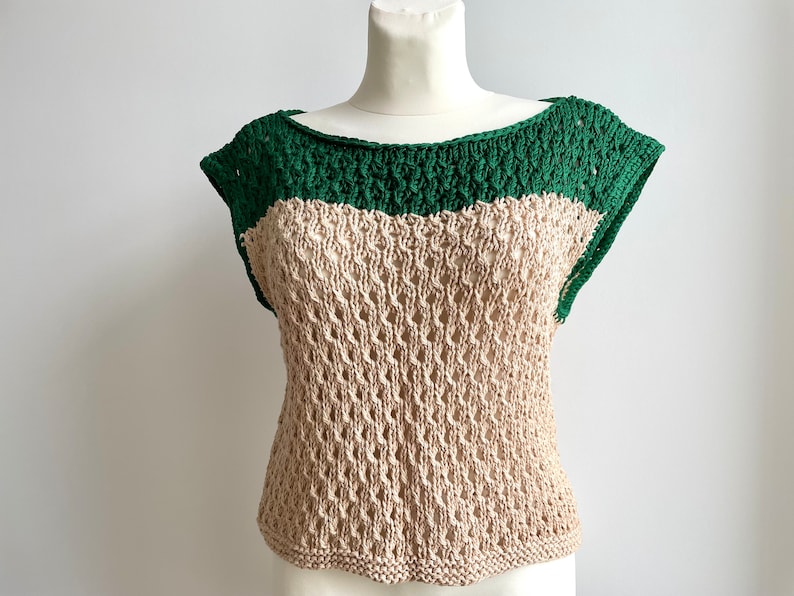 Puede incluir: Top de ganchillo hecho a mano, de dos tonos. El top presenta un escote y mangas cortas de color verde oscuro, con un cuerpo beige de tejido abierto. La prenda se exhibe en un maniqu&iacute;, mostrando su textura y dise&ntilde;o. Este top es una adici&oacute;n elegante a cualquier guardarropa.
