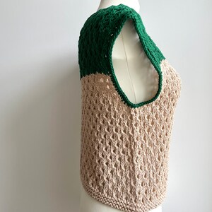 Puede incluir: Un top sin mangas de tejido abierto beige, con una secci&oacute;n superior y ribete verde oscuro. La prenda se exhibe en un maniqu&iacute;, destacando su textura y dise&ntilde;o. Presenta un escote redondo y un dobladillo redondeado. La prenda es de punto.