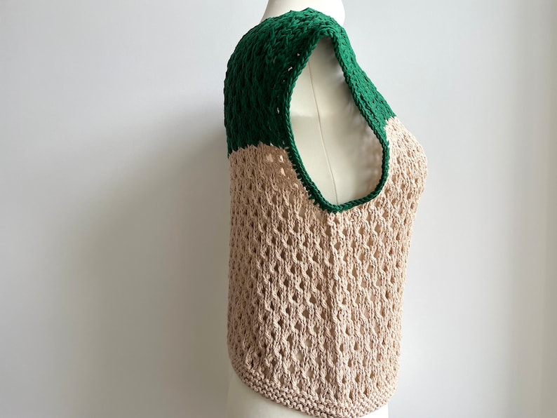 Puede incluir: Un top sin mangas de tejido abierto beige, con una secci&oacute;n superior y ribete verde oscuro. La prenda se exhibe en un maniqu&iacute;, destacando su textura y dise&ntilde;o. Presenta un escote redondo y un dobladillo redondeado. La prenda es de punto.