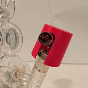 May include: A red glass pipe with a clear glass bowl and a red silicone mouthpiece. The pipe has a small, round, dark brown glass marble in the bowl.