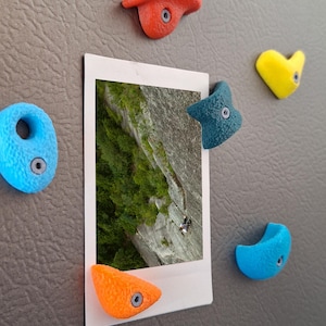 May include: A collection of colorful climbing hold magnets in red, blue, orange, and yellow surround a small photo of a rock climber on a cliff face. The magnets are attached to a gray surface.