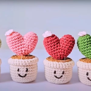 Könnte beinhalten: Gehäkelte herzförmige Kakteen in weißen Töpfen mit lächelnden Gesichtern. Die Herzen sind rosa, rot und grün, mit kleinen weißen Blumenakzenten. Die Töpfe haben einen hellbraunen Sockel.