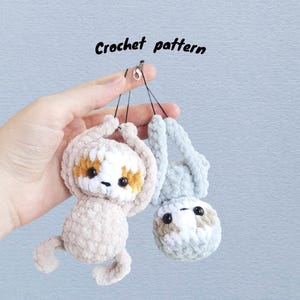 May include: Two crocheted sloth toys, one upright and one upside down. The sloths are beige and light blue with brown and white details. The text "Crochet pattern" is above the sloths. They are held by a hand.