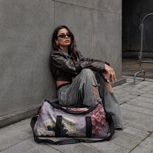 Puede incluir: Una bolsa de lona con estampado floral con correas negras y una correa para el hombro. La bolsa presenta un diseño con flores rosas, olas azules y un fondo morado. La bolsa está sobre una superficie gris.