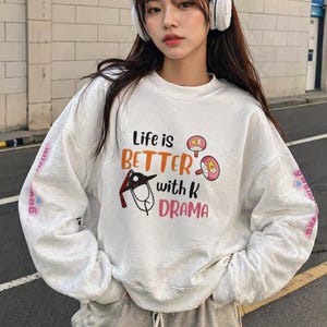 Puede incluir: Una sudadera blanca con el texto "Life is BETTER with K DRAMA" en un diseño colorido. La sudadera tiene mangas largas y cuello redondo. Las mangas tienen texto. La sudadera está hecha de un material suave.