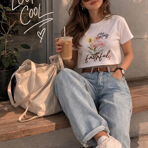 Puede incluir: Una mujer con un top blanco con flores y la frase "Stay Faithful", vaqueros claros y zapatillas blancas. Sostiene una bebida, con un bolso tote beige a su lado. La imagen tiene una estética cálida y soleada.