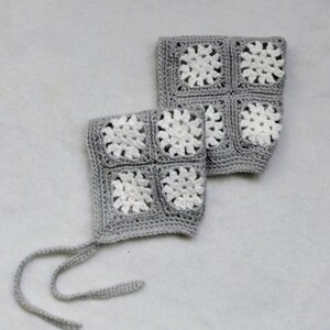 May include: Two crocheted baby bonnets. Each bonnet is gray with four white floral squares. One bonnet has two gray ties. The bonnets are handmade and likely for infants or toddlers. The bonnets are on a white surface.