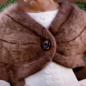 May include: A fuzzy, brown, knitted shrug featuring a textured, ribbed pattern and a single, large, dark, circular button closure at the center. The garment is worn over a white, long-sleeved, textured knit top with a subtle vertical cable pattern.