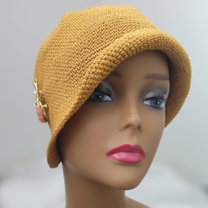 May include: Mustard yellow crochet cloche hat with a gold-tone and coral embellishment. The hat has an asymmetrical brim and a close-knit texture, offering a vintage aesthetic. A stylish head covering.