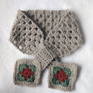 Op de afbeelding: Een handgemaakte gehaakte sjaal in een neutrale grijze kleur. De sjaal heeft een granny square design met twee vierkante zakken aan de uiteinden, elk met een rood en groen bloemmotief. De sjaal wordt op een witte ondergrond getoond.