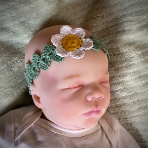 May include: A sleeping baby wearing a crocheted headband with a white flower and green leaves.