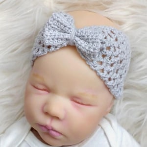 Könnte beinhalten: Ein graues Häkelband mit einer Schleife oben. Das Stirnband wird von einem Baby mit geschlossenen Augen getragen.