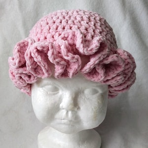 May include: A light pink, crocheted baby hat with a ruffled brim. The hat is made of a soft, plush material and is displayed on a white mannequin head. The hat's design features a textured crown and a decorative, wavy edge.