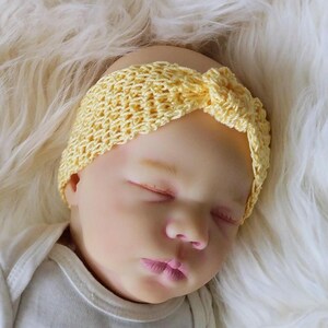 May include: A close-up of a baby wearing a yellow crocheted headband. The headband is knotted at the top. The baby is wearing a white onesie and is lying on a white, fluffy surface.
