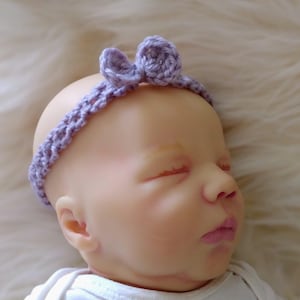 May include: A sleeping baby wearing a purple crocheted headband with a bow on top.