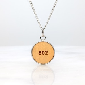 May include: A silver chain necklace with a round wooden pendant. The pendant has a light brown wood grain and features the number "802" in a dark brown font. The necklace is displayed on a white surface.