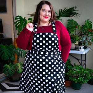 May include: A black and white polka dot apron with adjustable straps and pockets. The apron is worn over a long-sleeved burgundy shirt and black leggings. The person is wearing black shoes. The background includes green plants.
