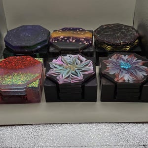May include: A set of six decorative coasters with unique designs. The coasters feature various colors, including red glitter, purple, gold, and floral patterns. Each coaster sits on a black square holder, offering a stylish and functional accessory.