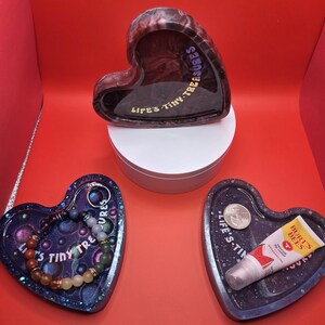 May include: Heart-shaped resin trays and a heart-shaped container. One tray holds a bracelet, another a tube of Burt's Bees lip balm and a coin. The container and trays have the text "LIFE'S TINY TREASURES".