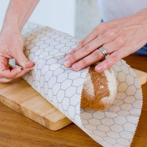 May include: A loaf of bread wrapped in white beeswax food wrap with a honeycomb pattern. The wrap is being folded over the bread by a hand.