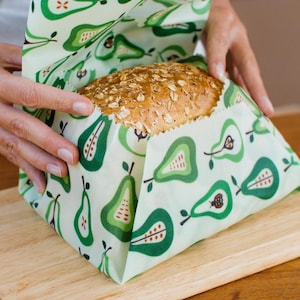 May include: A loaf of bread wrapped in green and white reusable food wrap with a pear pattern. The wrap is made of beeswax, cotton, and tree resin.