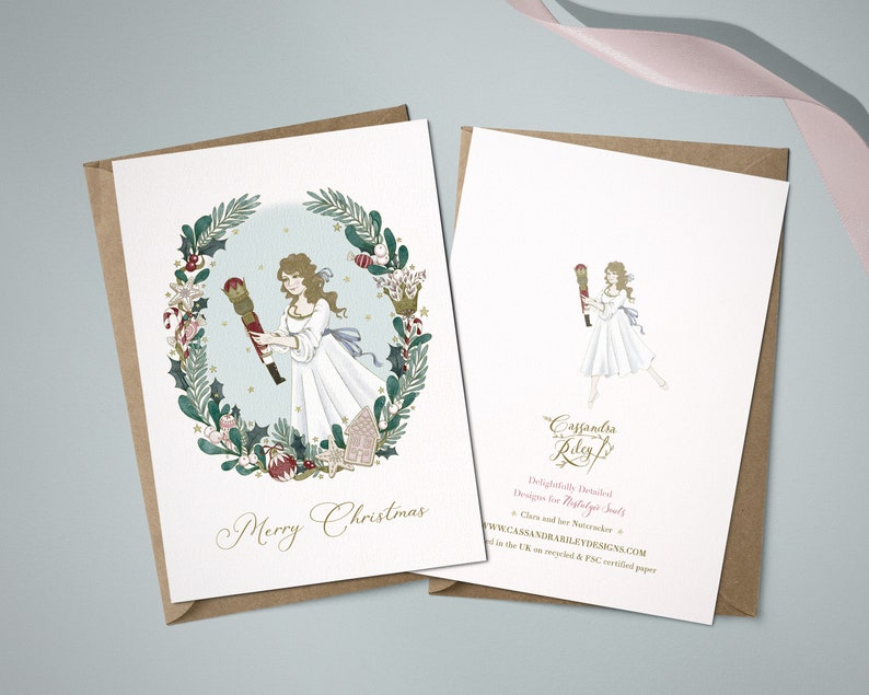 A Christmas card featuring a watercolour illustration of Clara in a white dress holding a nutcracker. The illustration is surrounded by a wreath of holly, berries, and other festive greenery. The card reads "Merry Christmas".
