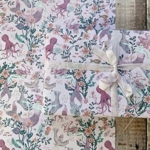 Pink wrapping paper with a repeating pattern of mermaids, octopuses, and sea life. The paper is wrapped around a gift box with a white ribbon.