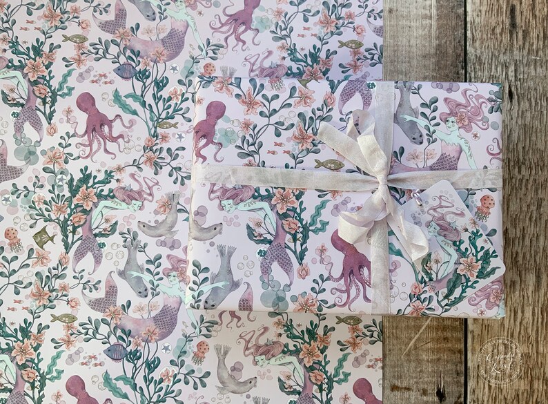 Pink wrapping paper with a repeating pattern of mermaids, octopuses, and sea life. The paper is wrapped around a gift box with a white ribbon.