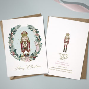 A Christmas card featuring a watercolour illustration of a Nutcracker in a red and gold uniform, standing in a wreath of greenery, candy canes, and other festive elements. The card reads "Merry Christmas".