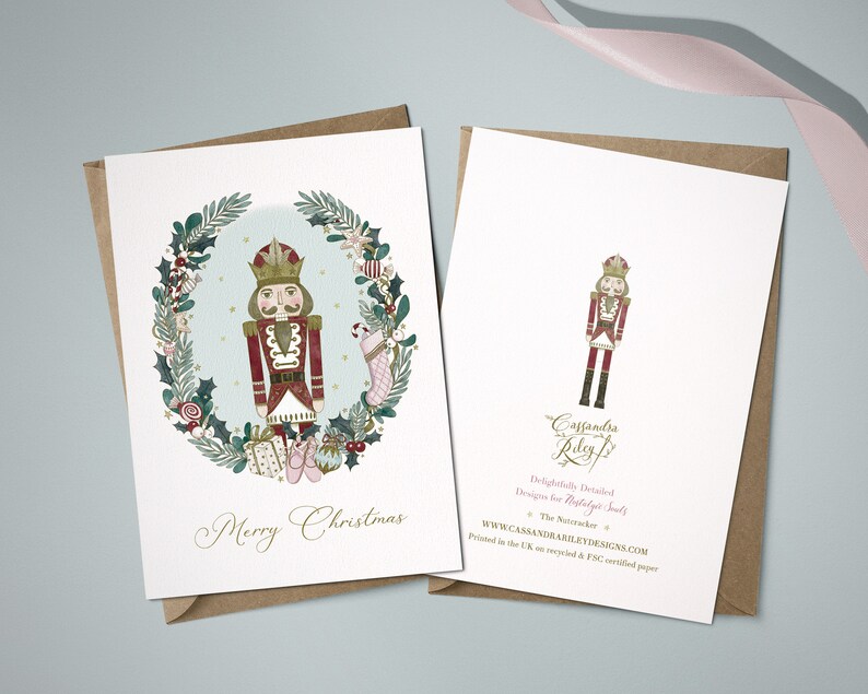 A Christmas card featuring a watercolour illustration of a Nutcracker in a red and gold uniform, standing in a wreath of greenery, candy canes, and other festive elements. The card reads "Merry Christmas".