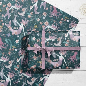 A sheet of teal blue wrapping paper with a repeating pattern of mermaids, octopuses, seashells, and flowers. The paper is wrapped around a gift box with a purple ribbon.