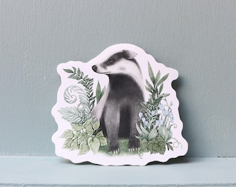 May include: A sticker featuring a detailed illustration of a badger with black and white fur, surrounded by green foliage and small blue flowers. The badger is sitting upright, with a serene expression.