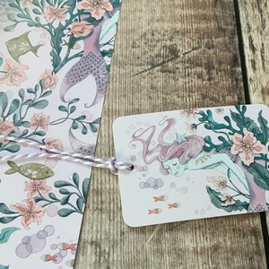 A gift tag featuring a pink and purple mermaid with a starfish in her hair is swimming in a sea of pink and green seaweed. The background is a light pink with white bubbles.