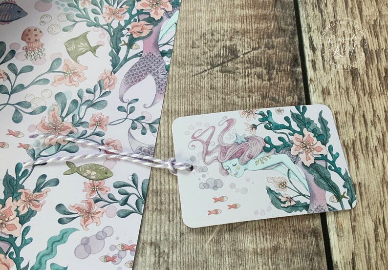 A gift tag featuring a pink and purple mermaid with a starfish in her hair is swimming in a sea of pink and green seaweed. The background is a light pink with white bubbles.