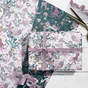 Two sheets of wrapping paper featuring mermaids, octopus, seals and sea life design. One sheet has a pink background and the other has a dark teal background. A gift box is wrapped with the pink wrapping paper and tied with a pink ribbon.