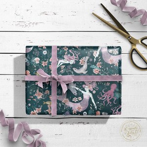 Teal blue gift wrap with a pink ribbon. The wrapping paper features a repeating pattern of mermaids, sea creatures, and flowers. The text "Casandra Riley" is visible in the lower right corner.