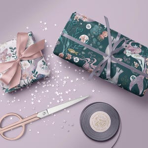 Two gift boxes wrapped in mermaid-themed wrapping paper. One box is wrapped in paper with pink and blue mermaids and sea creatures. The other box is wrapped in dark teal paper with pink mermaids and sea creatures. Both boxes are tied with pink ribbon. The boxes are on a light purple surface with silver glitter stars scattered around them.