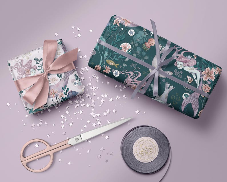 Two gift boxes wrapped in mermaid-themed wrapping paper. One box is wrapped in paper with pink and blue mermaids and sea creatures. The other box is wrapped in dark teal paper with pink mermaids and sea creatures. Both boxes are tied with pink ribbon. The boxes are on a light purple surface with silver glitter stars scattered around them.