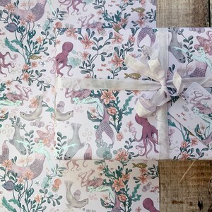 Pink wrapping paper featuring a mermaid, octopus, and sea life design. The paper is wrapped around a gift box with a white ribbon and a gift tag.