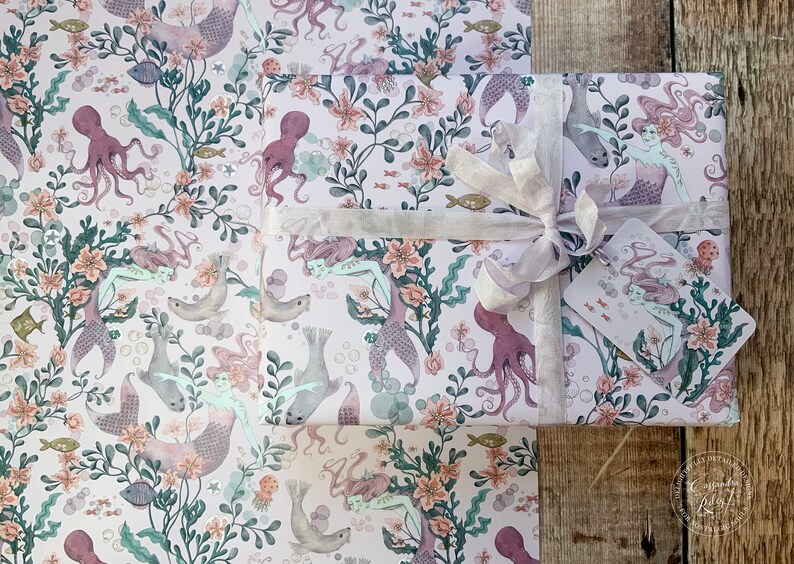 Pink wrapping paper featuring a mermaid, octopus, and sea life design. The paper is wrapped around a gift box with a white ribbon and a gift tag.