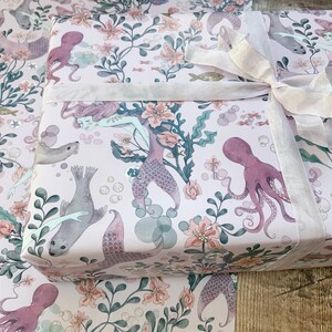 Pink wrapping paper featuring a mermaid, octopus, and sea life design. The paper is wrapped around a gift box with a white satin ribbon.