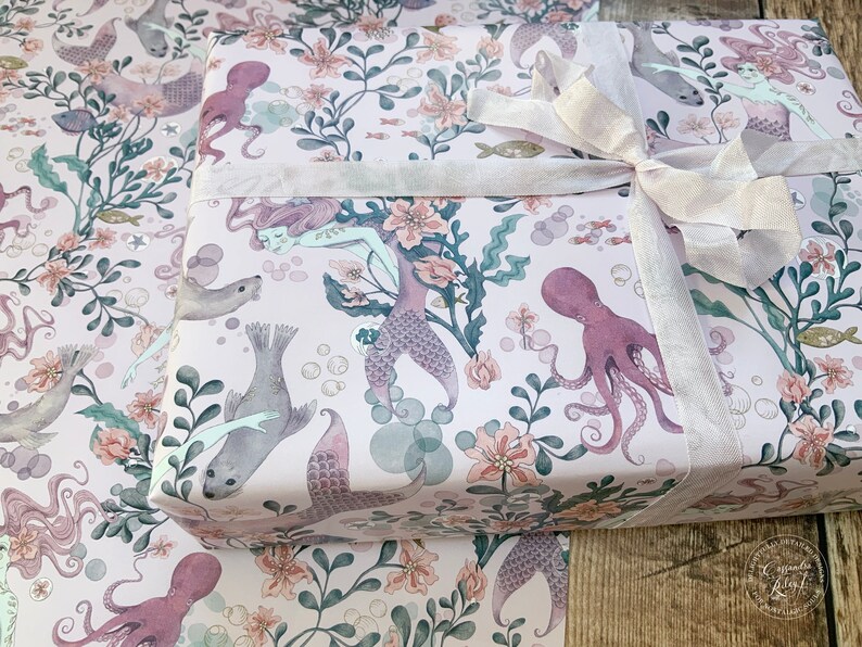 Pink wrapping paper featuring a mermaid, octopus, and sea life design. The paper is wrapped around a gift box with a white satin ribbon.