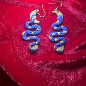 Puede incluir: Un par de pendientes de serpiente azul y dorado con un diseño de media luna. Los pendientes están hechos de arcilla polimérica y tienen un diseño colgante.