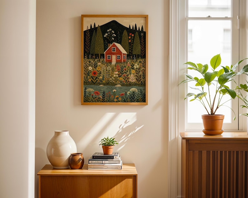 May include: A framed art print depicts a red house nestled in a field of flowers, with a mountain backdrop. The artwork is in a wooden frame and hangs on a cream-colored wall. A wooden cabinet with vases and books is in the foreground.